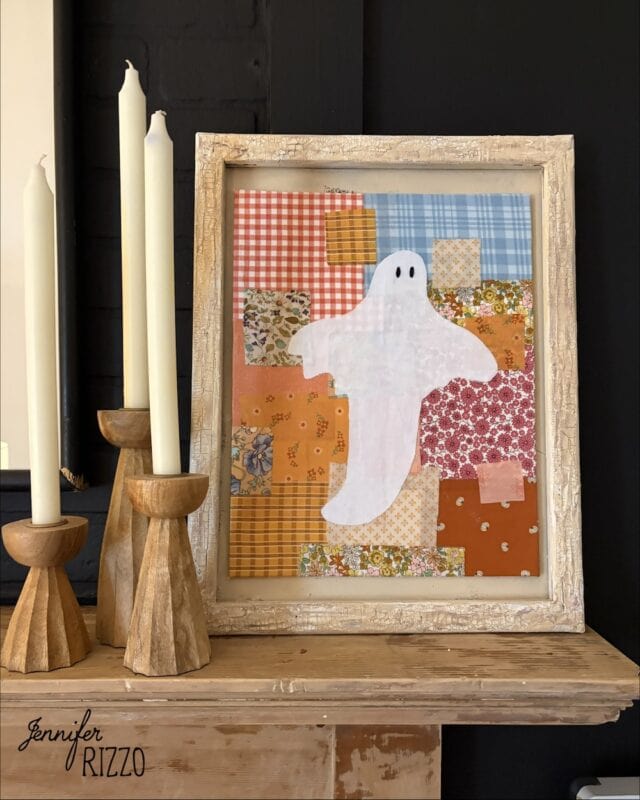 Framed patchwork art featuring a white ghost on colorful fabric squares, displayed on a mantel next to three wooden candle holders with tall candles.