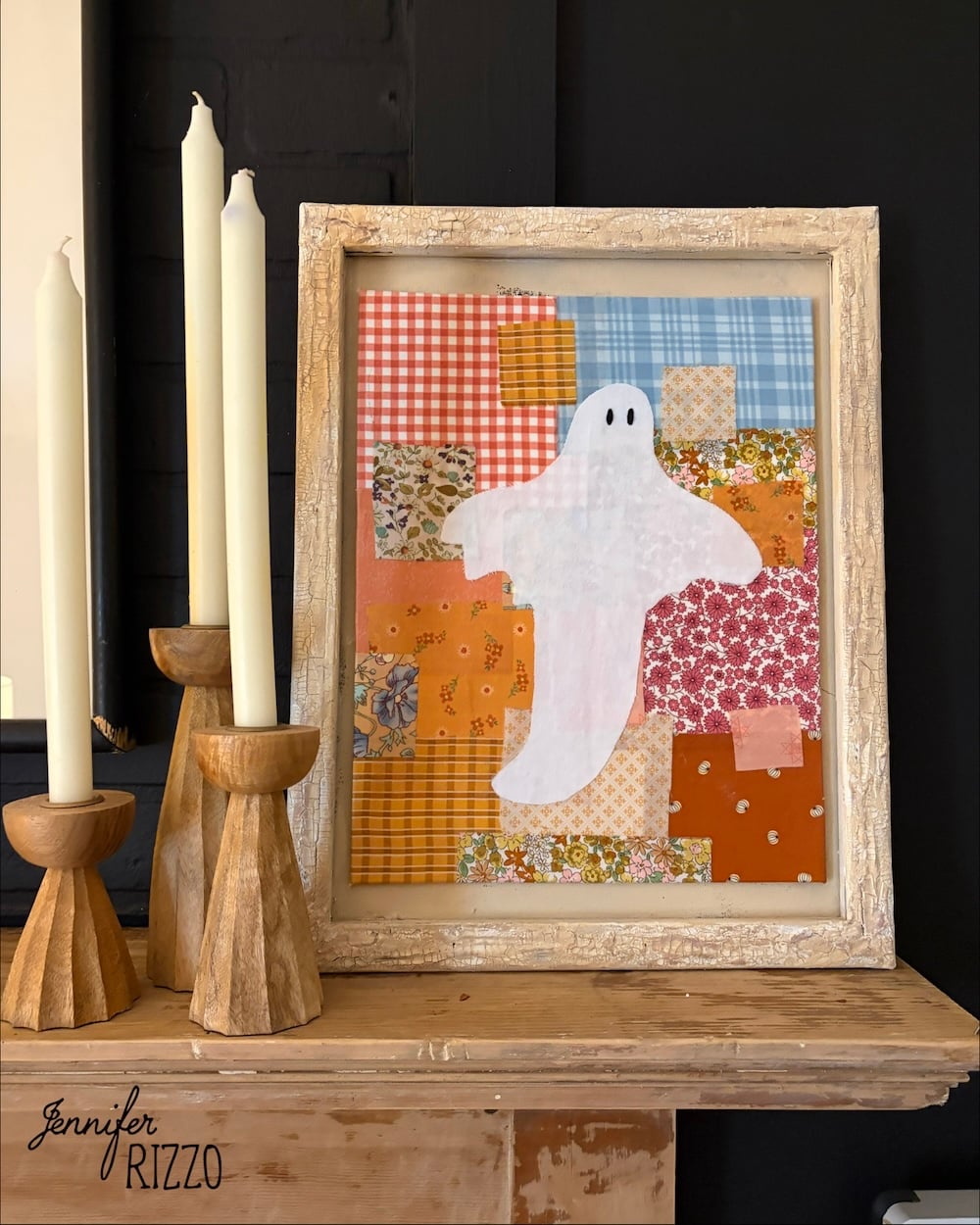Framed patchwork art featuring a white ghost on colorful fabric squares, displayed on a mantel next to three wooden candle holders with tall candles.