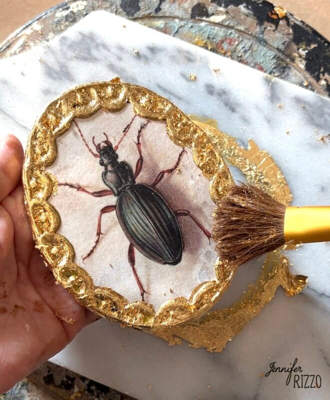 A hand holds an oval frame with a beetle illustration, as a brush applies gold leaf to the scalloped border.