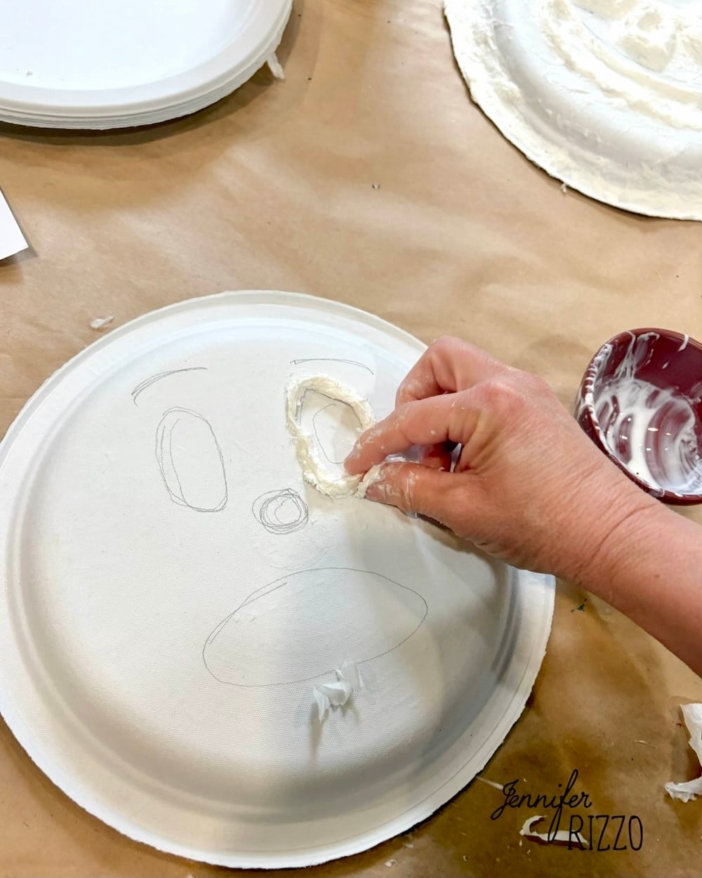 A hand adds a plaster piece to a paper plate with a face sketched on it, creating a 3D facial feature on the craft project.