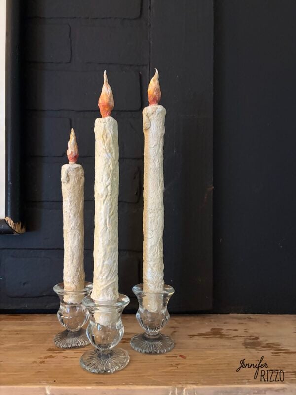 Three tall, textured white candles with faux flames stand in clear glass candle holders on a wooden surface against a dark background.