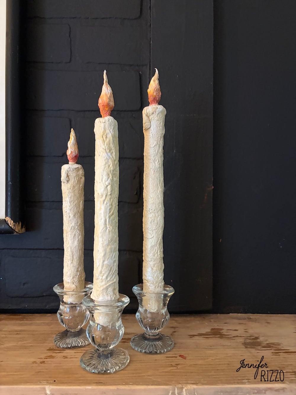 Three tall, textured white candles with faux flames stand in clear glass candle holders on a wooden surface against a dark background.