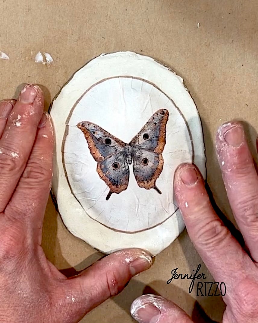 Two hands with paint on the fingers hold an oval wood slice featuring an image of a brown moth or butterfly centered on a light background.