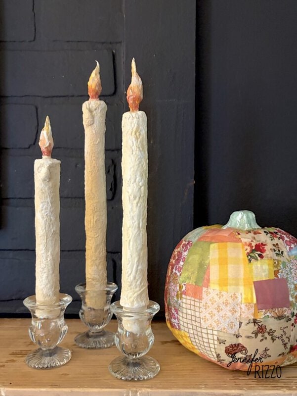 Three cream-colored candles in glass holders are displayed on a wooden surface next to a patchwork fabric pumpkin against a dark background.