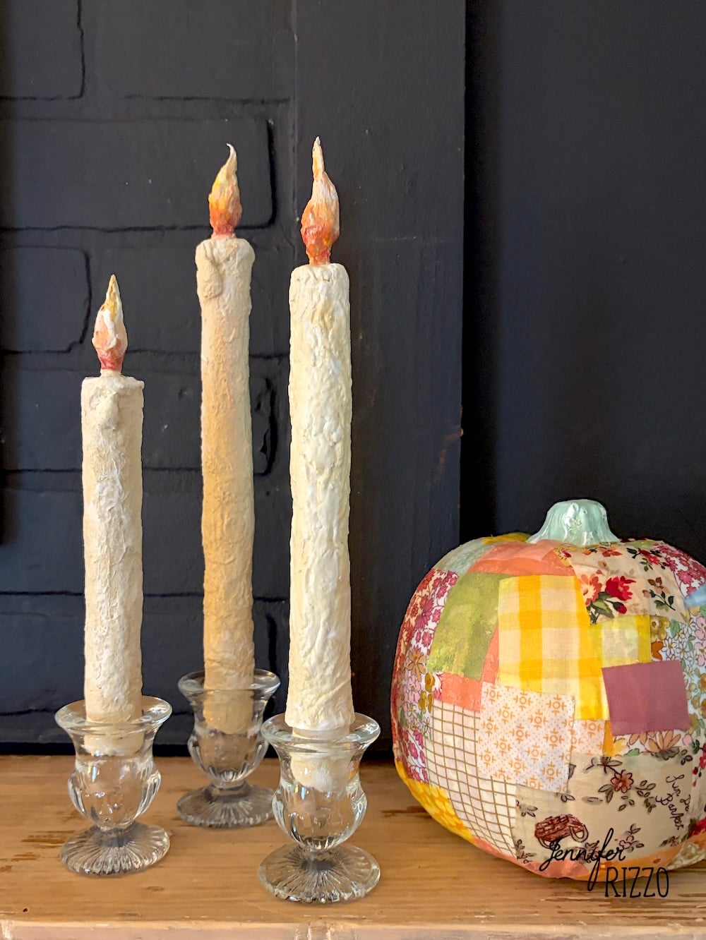 Three cream-colored candles in glass holders are displayed on a wooden surface next to a patchwork fabric pumpkin against a dark background.