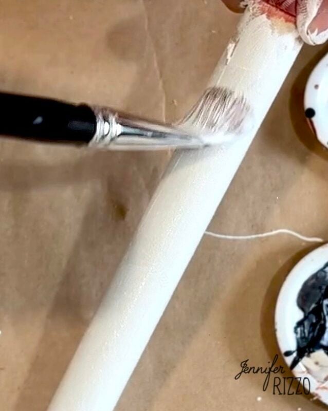 A person uses a paintbrush to apply white paint to a cylindrical object on a brown surface, with paint palettes nearby.
