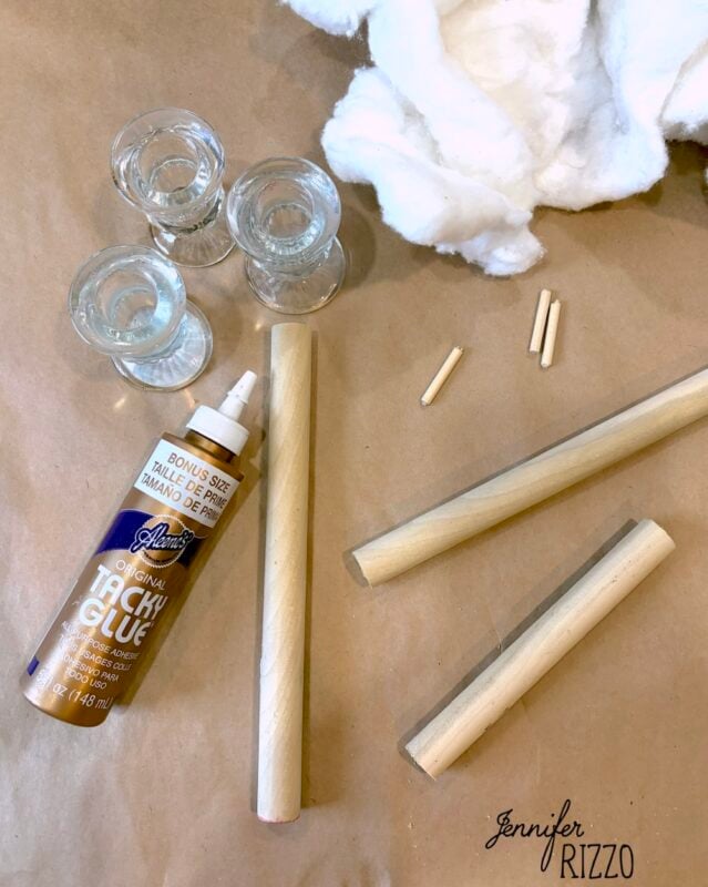 Three glass candle holders, cotton, tacky glue, wooden dowels, and dowel pieces are arranged on a brown surface.