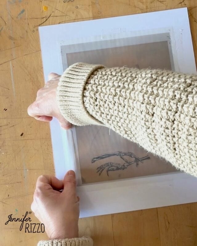 A person in a knitted sweater holds a screen over a paper with a hand skeleton drawing on a wooden table.
