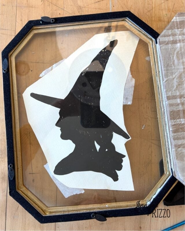 A glass panel with the black silhouette of a witch in a pointed hat, viewed from the side, placed on a wooden surface.