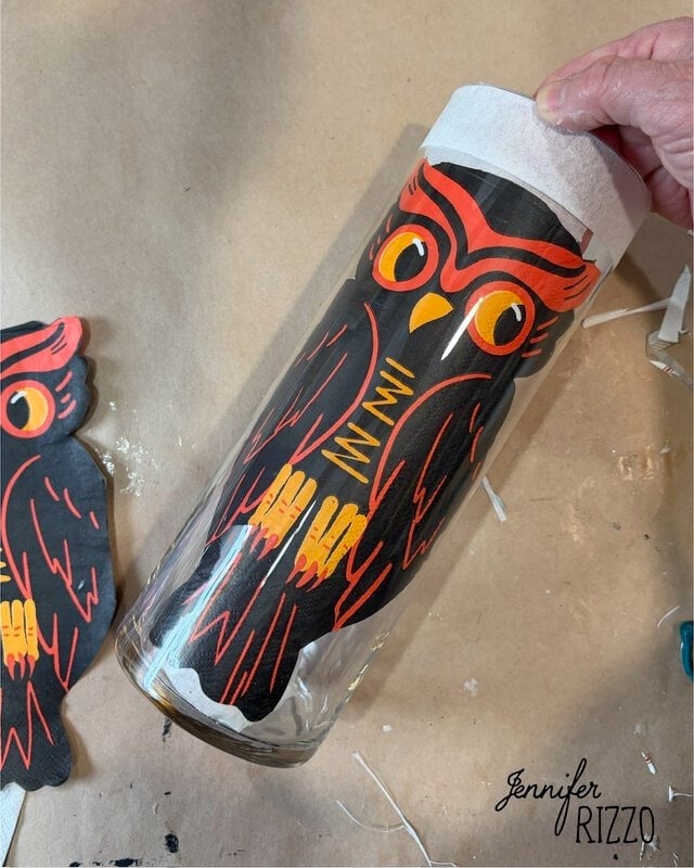 A hand applies a vintage-style black and orange owl decal to a clear glass vase on a beige craft surface.