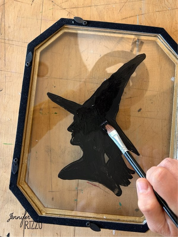 A hand paints a black silhouette of a witch with a pointed hat on the glass of an octagonal picture frame, resting on a wooden surface.