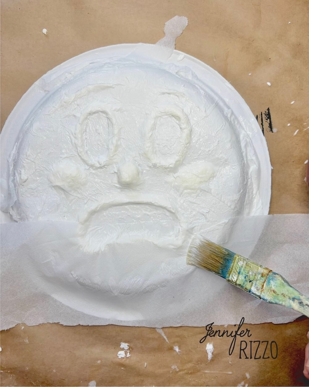 A paintbrush applies white paint to a paper plate with a sad face molded into its surface.