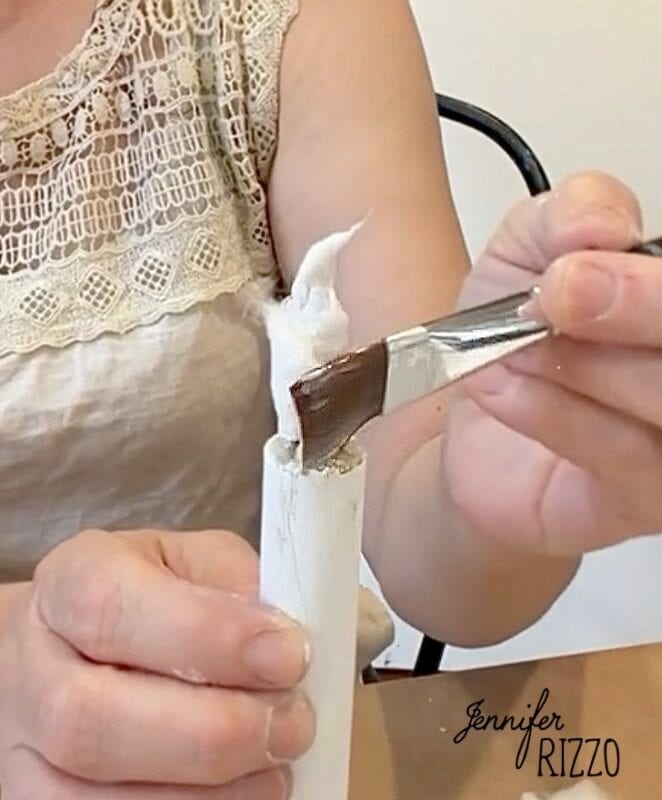 Person applies brown paint with a brush to the end of a white cardboard tube wrapped in cloth. The watermark “Jennifer Rizzo” is in the lower right corner.