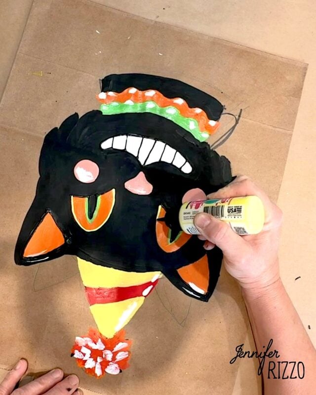 A person decorates a painted black cat face with orange eyes, party hat, and toothy grin on cardboard using yellow paint. “Jennifer Rizzo” is printed in the lower right corner.
