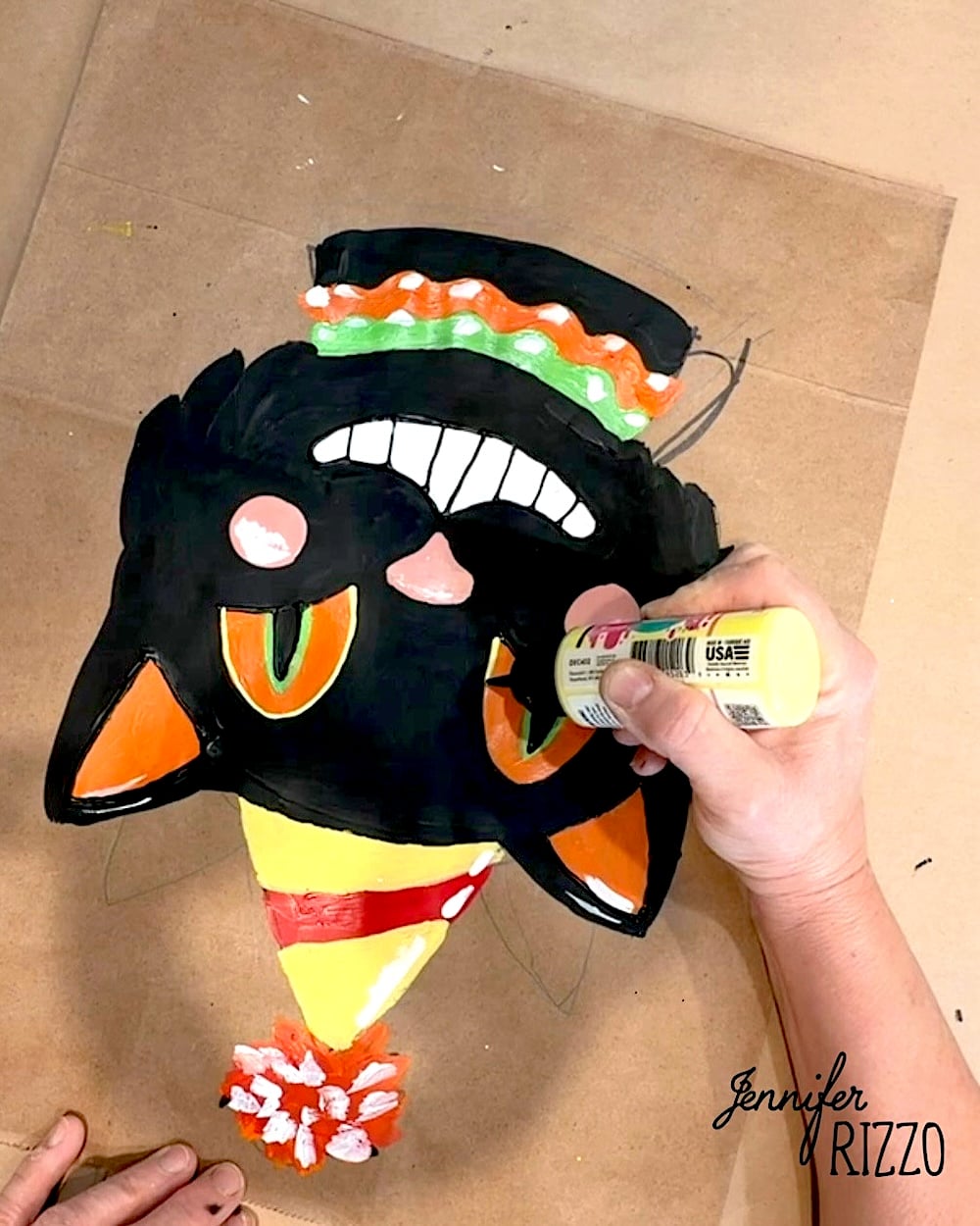 A person decorates a painted black cat face with orange eyes, party hat, and toothy grin on cardboard using yellow paint. “Jennifer Rizzo” is printed in the lower right corner.