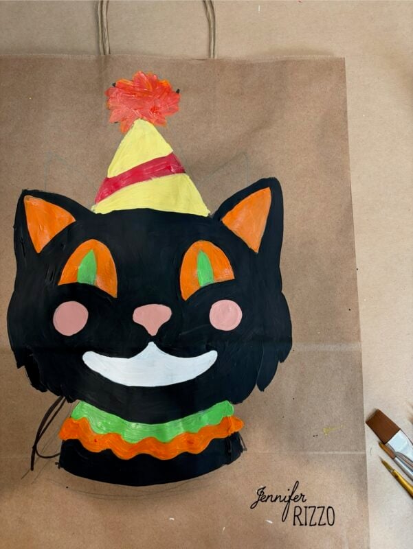 A painted black cat with orange eyes and a party hat on a brown paper bag. Paintbrush and signature “Jennifer Rizzo” are visible nearby.