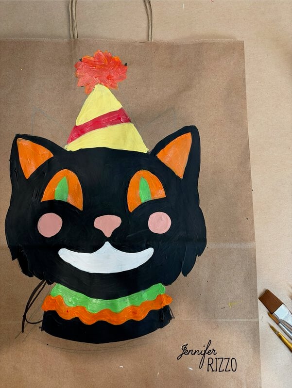 A painted black cat with orange eyes and a party hat on a brown paper bag. Paintbrush and signature “Jennifer Rizzo” are visible nearby.