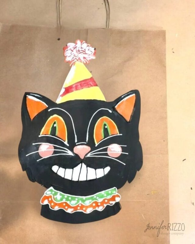 A painted black cat face with a party hat and ruffled collar is displayed on a brown paper bag.