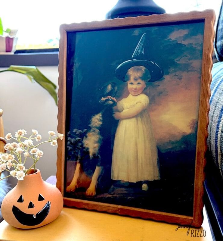 Framed painting of a child in a white dress and black witch hat standing beside a dog, placed on a table next to a small jack-o’-lantern vase with white flowers.