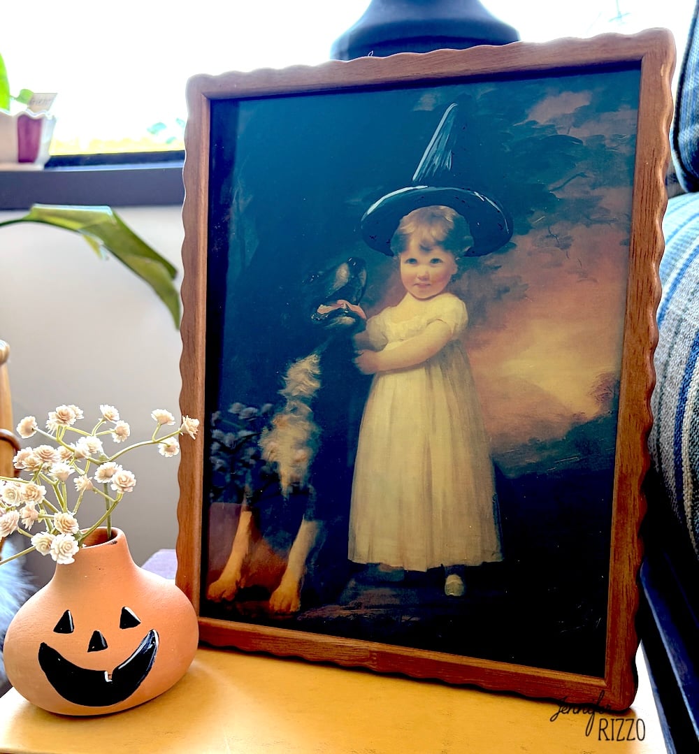 Framed painting of a child in a white dress and black witch hat standing beside a dog, placed on a table next to a small jack-o’-lantern vase with white flowers.