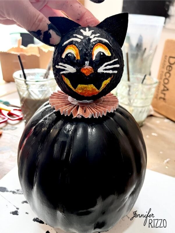 A black cat head with painted orange and white features sits atop a black pumpkin, with a pink frilled collar at the base. Art supplies and paint brushes are visible in the background.