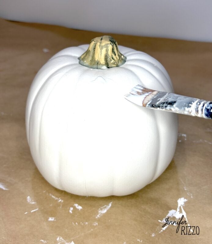 A paintbrush applying white paint to a small pumpkin with a gold-painted stem.