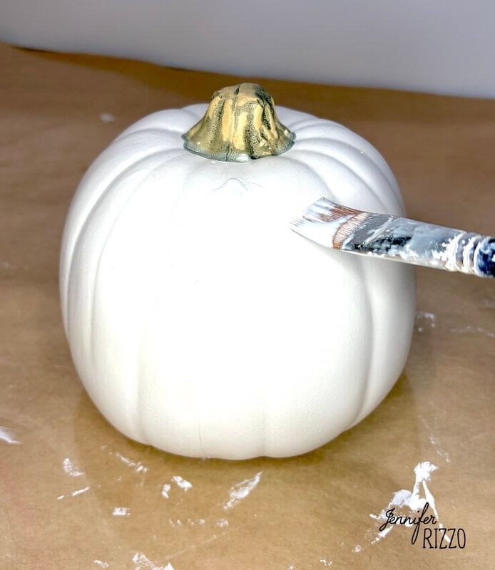 A paintbrush applying white paint to a small pumpkin with a gold-painted stem.