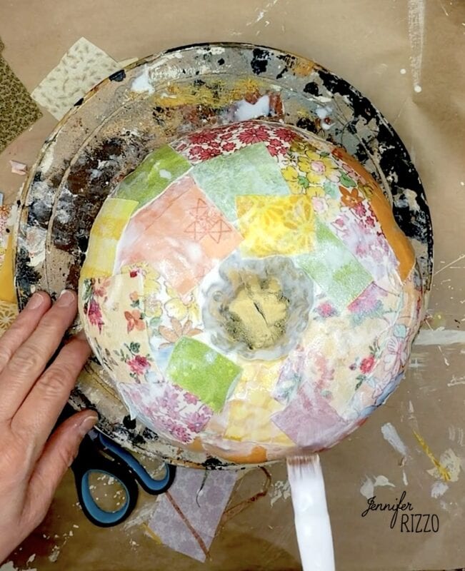 A person decoupaging colorful patterned paper onto a round bowl on a pottery wheel.