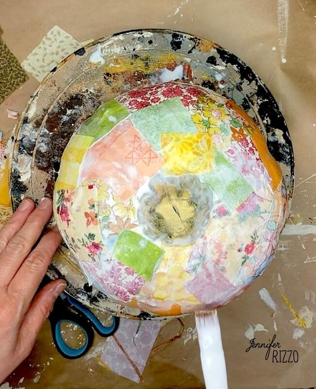 A person decoupaging colorful patterned paper onto a round bowl on a pottery wheel.