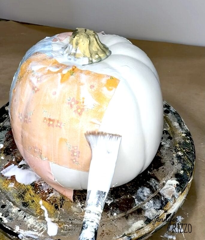 A paintbrush applies decoupage to a white pumpkin partially covered in floral patterned paper.