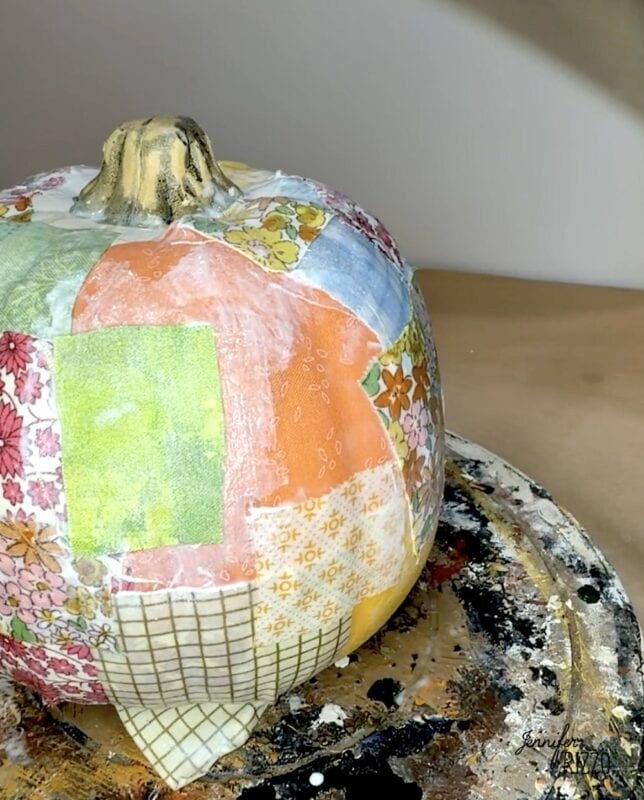A pumpkin covered in patchwork fabric squares, sitting on a paint-splattered surface.
