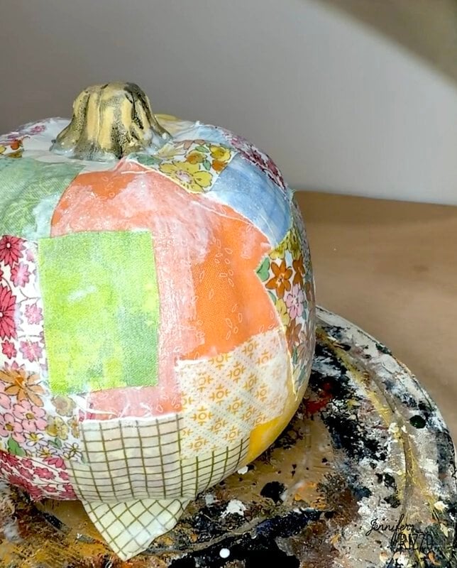 A pumpkin covered in patchwork fabric squares, sitting on a paint-splattered surface.