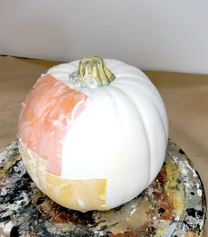 A white pumpkin partially covered with pieces of gold leaf, sitting on a weathered surface.