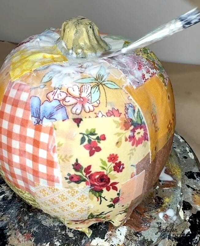 A paintbrush applies decoupage glue to a pumpkin covered in patches of floral and checkered fabric.