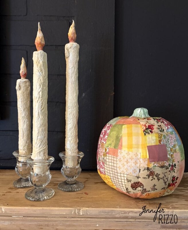 Three textured taper candles in clear glass holders next to a pumpkin covered in a patchwork of patterned fabric.