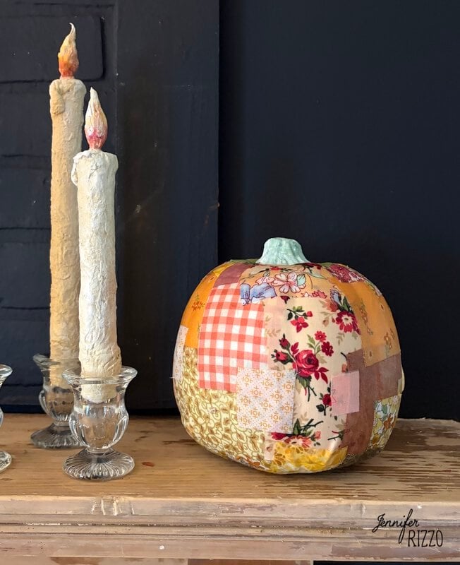 A fabric-covered pumpkin decorated with assorted floral and checkered patches next to two tall taper candles in glass holders.
